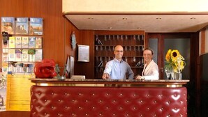 Ein Empfangsschalter mit roter Polsterung, dahinter ein Mann und eine Frau sowie eine Wand mit gerahmten Bildern und einem Aushangbrett.