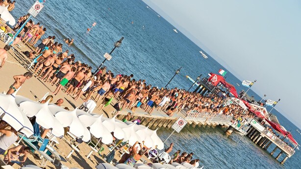 Ein überfüllter Strand mit vielen weißen Liegestühlen und einer Mole, die sich ins Wasser erstreckt.
