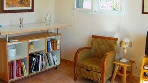Ein Wohnzimmer mit Bücherregal, einem Korbstuhl, einem kleinen Tisch mit Lampe und einem gerahmten Bild an der Wand.