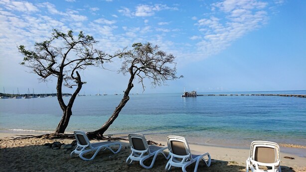 Une plage avec des chaises longues blanches, un ciel dégagé et une mer calme où l’on aperçoit des bateaux au loin.