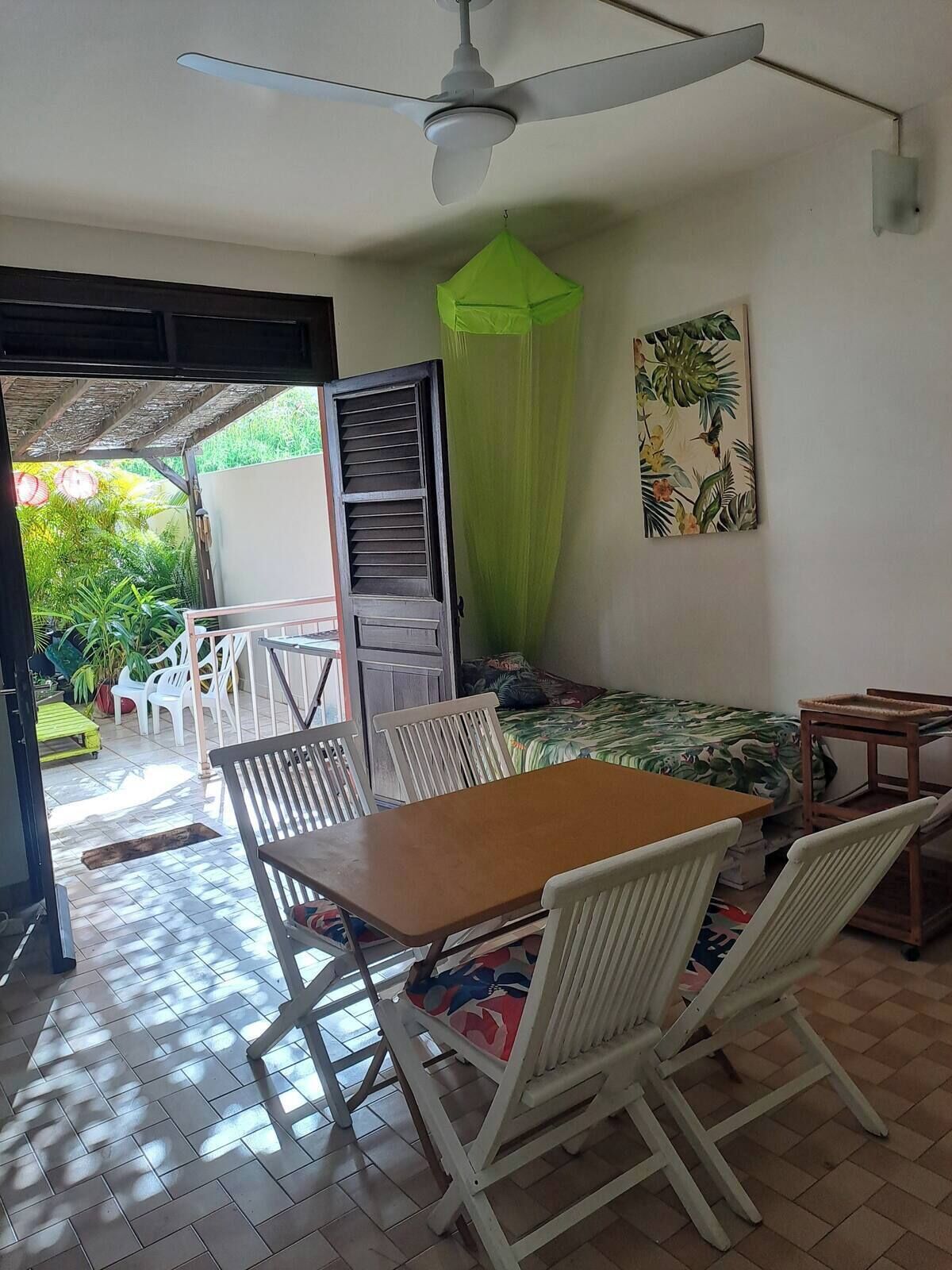 Una habitación con una mesa de comedor, sillas, un ventilador de techo, una cama con dosel verde y un cuadro de plantas tropicales.