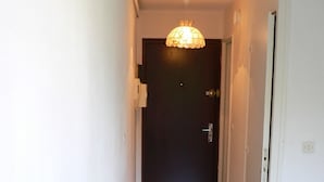 Un couloir étroit avec une table blanche et deux chaises, une porte sombre au fond, et un ventilateur de plafond.