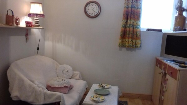 A cozy living room with a white armchair, a small table with plates, a clock on the wall, and a television on a wooden cabinet.