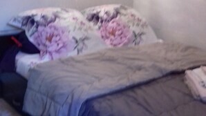 Un dormitori amb un llit cobert per un llençol amb estampat floral, un edredó gris i un almoiner gris.