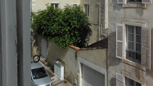 A narrow street with buildings on either side, a parked car, and a tree with green foliage.