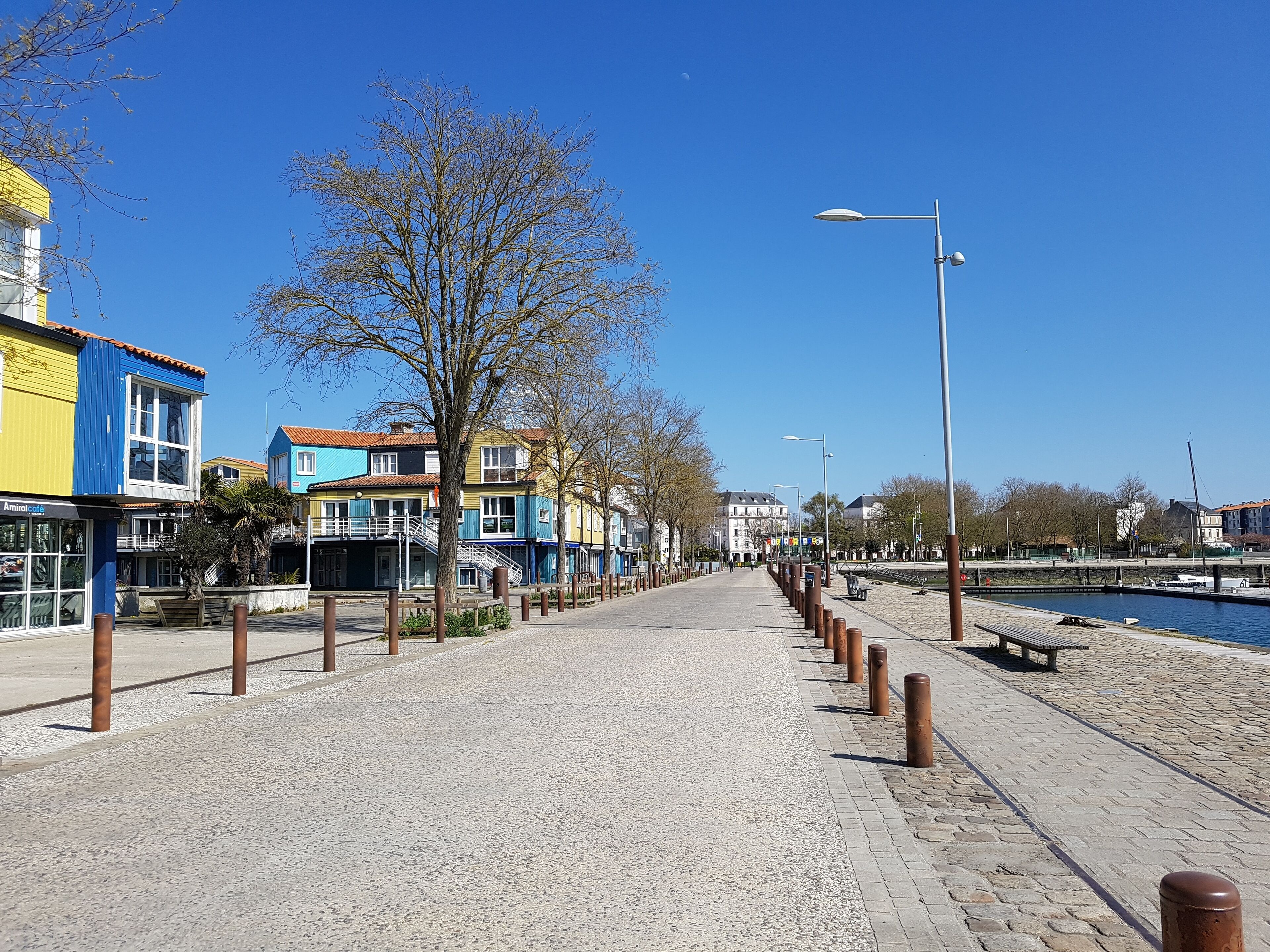Ein gepflasterter Weg entlang eines Gewässers, gesäumt von Leitpfosten und Bäumen, der zu einer Reihe bunter Gebäude führt.