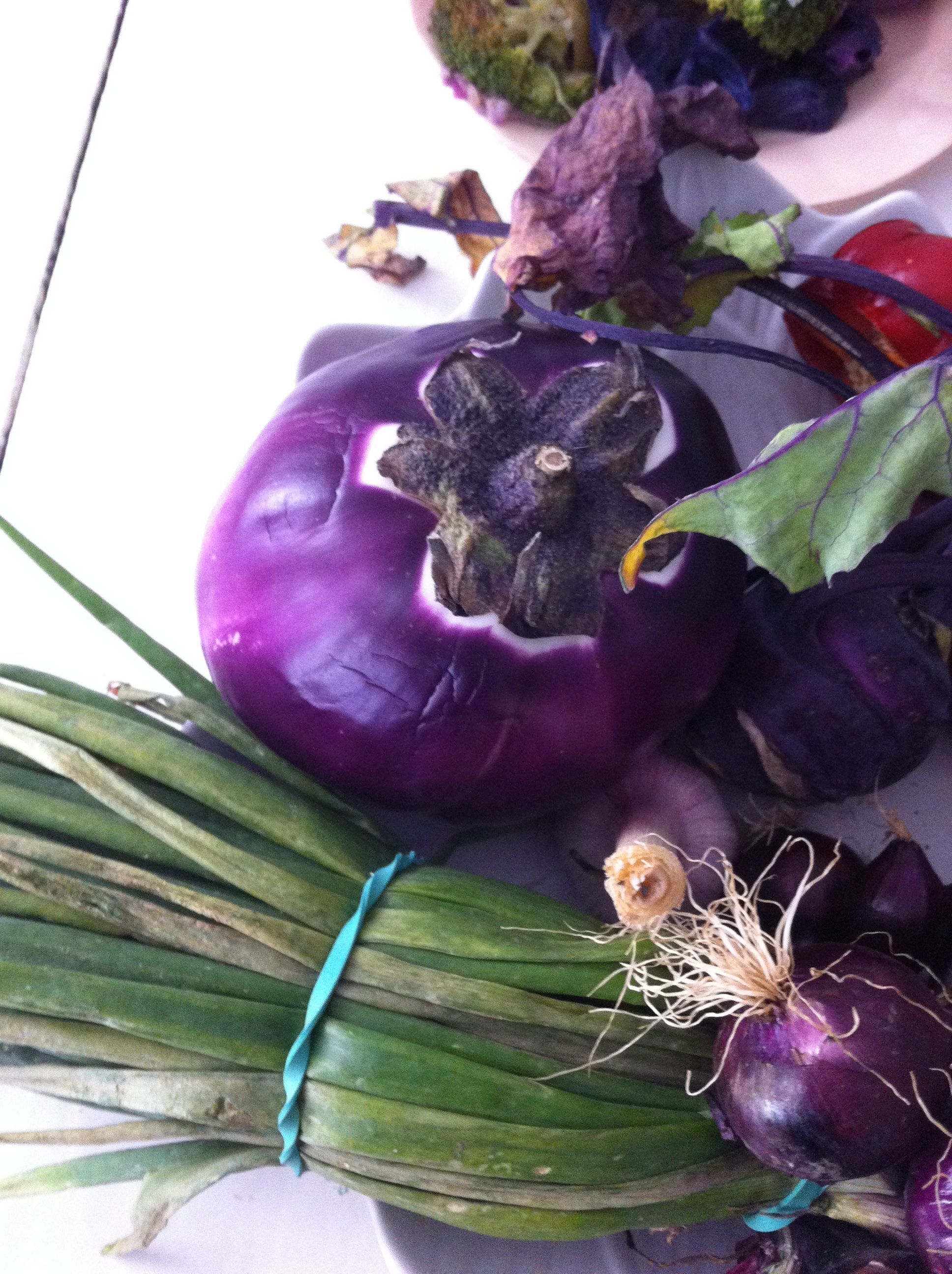 A purple eggplant, green onions, and a red bell pepper on a white surface.