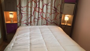 Una cama bien hecha con edredón blanco, flanqueada por dos mesitas de noche con lámparas, en una habitación con cortinas estampadas y una pared de acento morada.