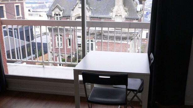 Une pièce avec une table blanche et une chaise noire, une fenêtre donnant sur des immeubles et un plancher en bois.