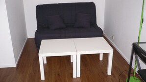Une pièce avec un canapé de couleur sombre placé contre un mur blanc, deux tables blanches au centre et un lampadaire vert sur la droite.