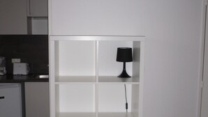 Une étagère blanche surmontée d’une lampe noire, située dans une pièce dont on aperçoit un coin cuisine au fond.