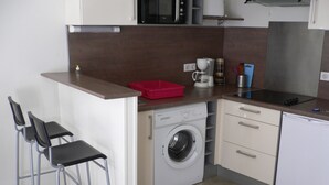 A compact kitchen with a washing machine, a coffee maker, and a microwave. There are two bar stools and a red container on the counter. - Historic center - Old market (Rouen)