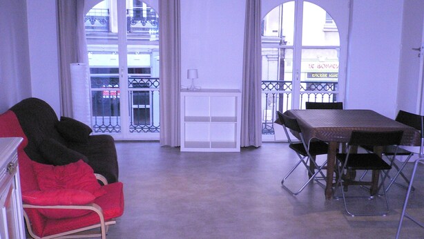 A room with a red chair, a table with chairs, and a view of a street through large windows. - Historic center - Old market (Rouen)