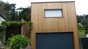 Un edificio moderno in legno con un'auto in garage, immerso nel verde e circondato da una recinzione in legno.
