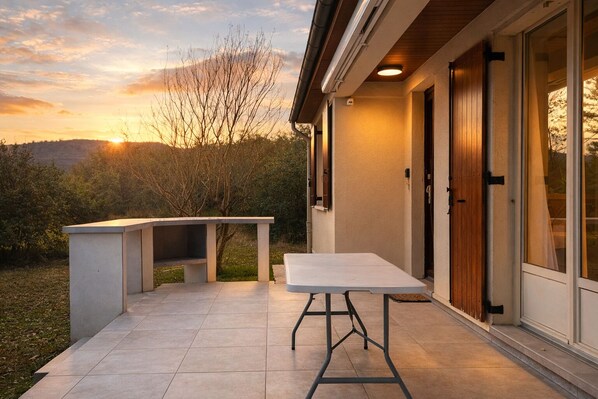 Um pátio externo com mesa, banco, uma porta de madeira e vista para o pôr do sol.