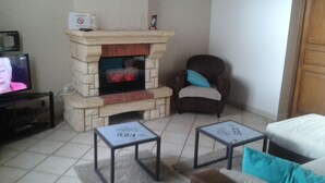 Ein Wohnzimmer mit Kamin aus Stein, einem Fernseher, zwei kleinen Tischen und einem braunen Sessel mit blauem Kissen.