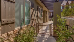 A covered outdoor area with a stone wall, a metal gate, and a pathway leading to a building with large windows and a pergola.