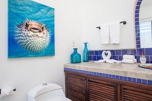 A bathroom with a pufferfish painting, a toilet, a sink with blue tiles, and a mirror with a blue frame. - BEAUTIFUL VILLA WITH PRIVATE POOL (Cabo San Lucas)