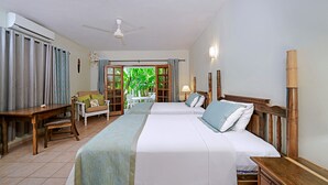 Une chambre d’hôtel avec deux lits, un bureau en bois, une chaise, un ventilateur de plafond et une vue sur la verdure environnante.