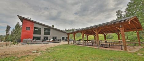 Un edificio moderno con fachada roja y gris, un área de picnic techada con bancos y un campo de césped en primer plano.