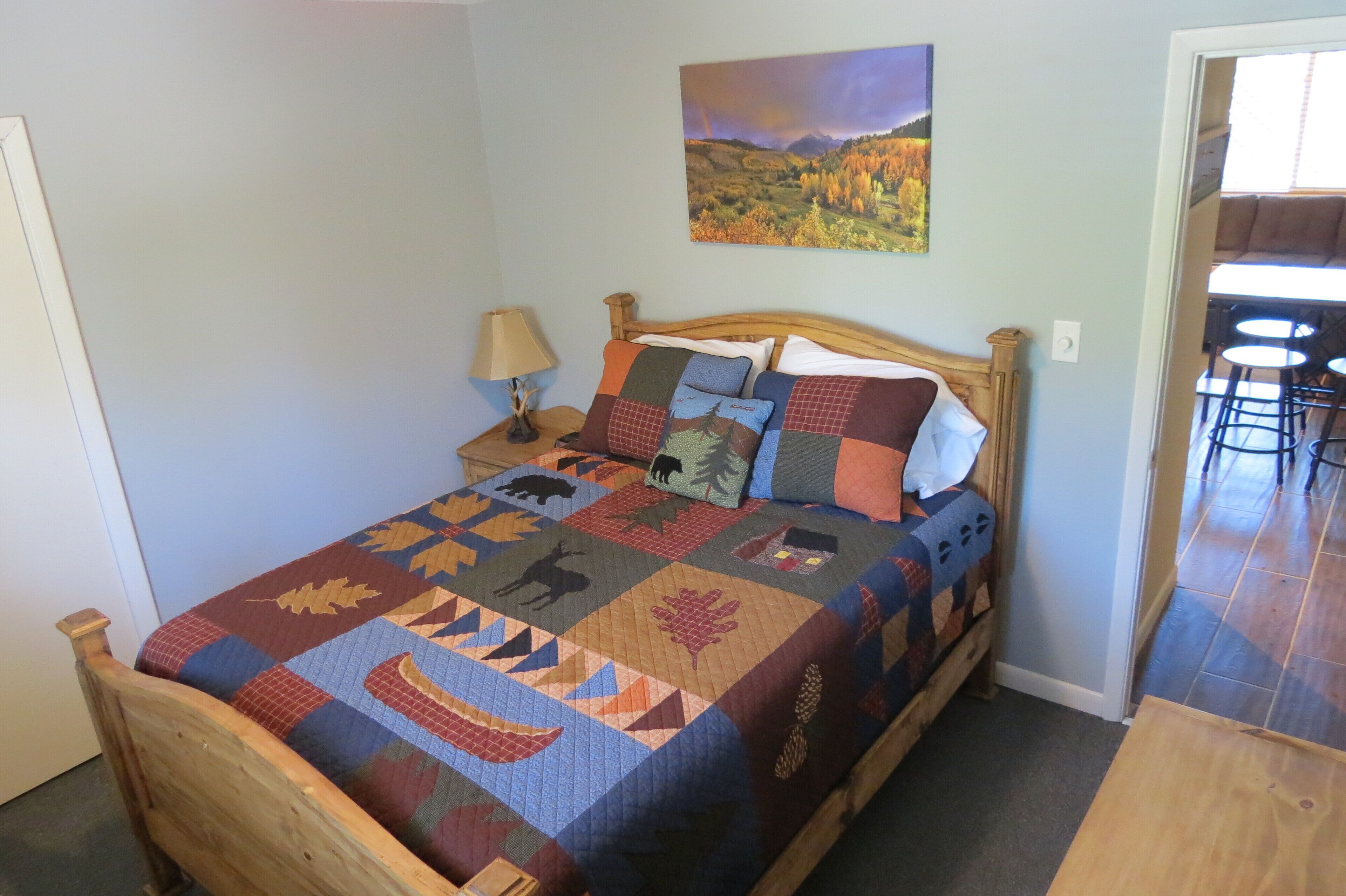 Een bed met een kleurrijke quilt, een nachtkastje met een lamp, en een schilderij van een landschap aan de muur.