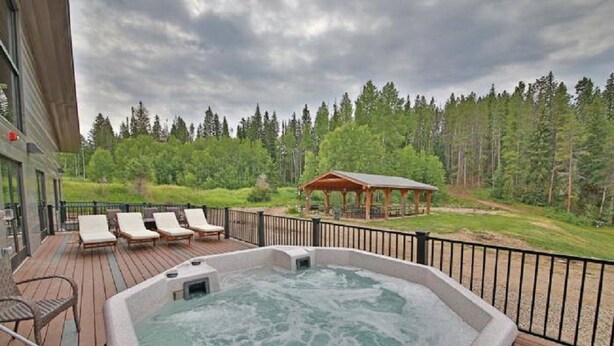 Un jacuzzi en una terraza de madera con sillones reclinables, un pabellón y un paisaje boscoso de fondo.