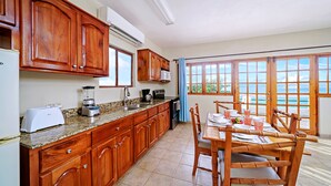 A kitchen with wooden cabinets, a granite countertop, and a dining area with a table and chairs. - Beach House Condos: Beachfront Penthouse - 7mile Beach Jamaica (Negril)