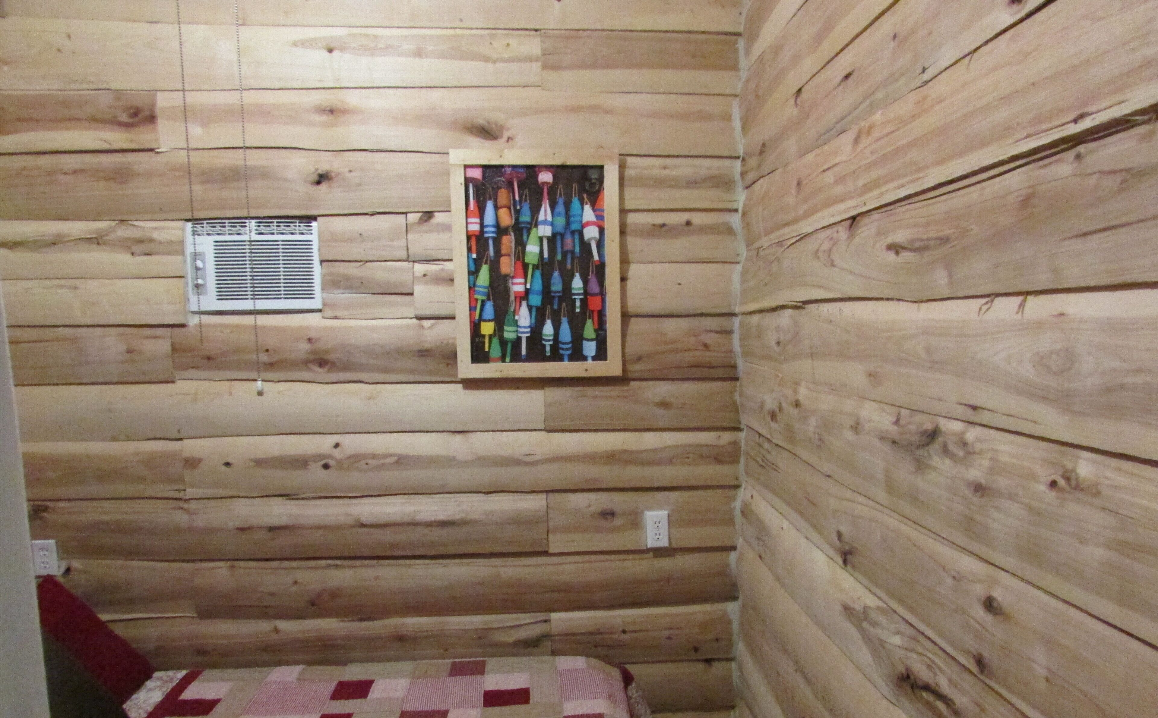 A wooden cabin interior with a quilted bed, a wall-mounted air vent, and a framed picture of colorful buoys.