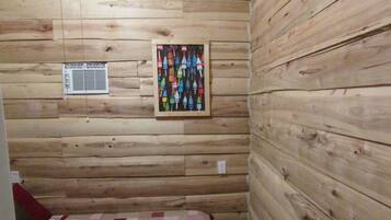 A wooden cabin interior with a quilted bed, a wall-mounted air vent, and a framed picture of colorful buoys.