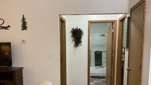 Un salon avec un canapé blanc, une table basse en bois et un téléviseur posé sur un meuble. Un couloir mène à une salle de bain équipée d’une douche et où une serviette est accrochée à la porte.