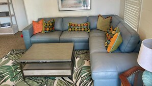 Ein Wohnzimmer mit grauem Sofa, bunten Kissen mit Muster, einem Holztisch und einem gerahmten Kunstwerk an der Wand, sowie einem Fenster mit Jalousien.