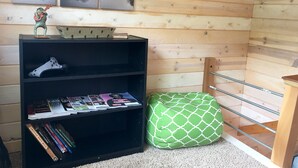 A room with a wooden sloped ceiling, a black bookshelf with magazines, a green patterned bean bag, and two framed pictures on the wall. - Red Tiny House In The Mountains - waterfall, jacuzzi and stunning farm views (Clayton)