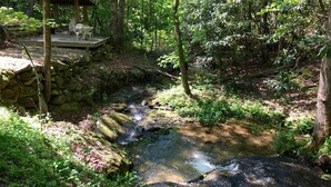 A rustic wooden deck with chairs, a stone wall, and a small wooden structure in a forested area with a stream. - Red Tiny House In The Mountains - waterfall, jacuzzi and stunning farm views (Clayton)