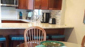 Een keuken met houten kastjes, een roestvrijstalen magnetron, een koffiezetapparaat en een broodrooster. Er staat een glazen tafel met blauwe stoelen en een decoratieve schaal erop.