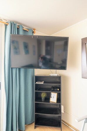 A corner room with a flat-screen TV mounted on the wall, a black entertainment unit with a plant and framed certificate, and teal curtains. - Need Vitamin Sea??? 2br/2baths (San Miguel)