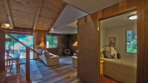 A cozy living room with a fireplace, a dining area with a table and chairs, and a view of the outdoors through a large window. - The Lyons Den at Upper Timberline Village (Packwood)
