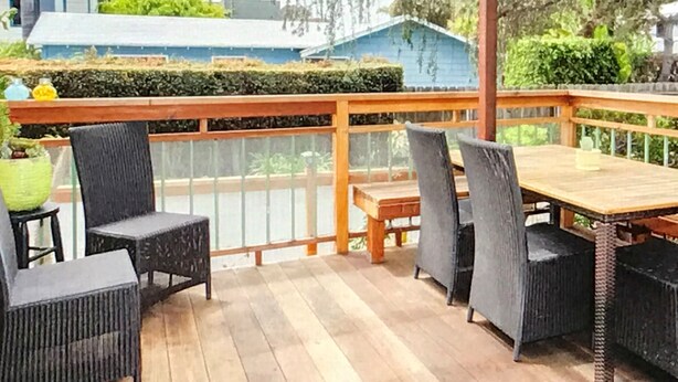 A wooden deck with a table and chairs, surrounded by greenery and a blue building in the background. - Cozy 2-Bedroom Home Just Blocks from the Beach (Encinitas)