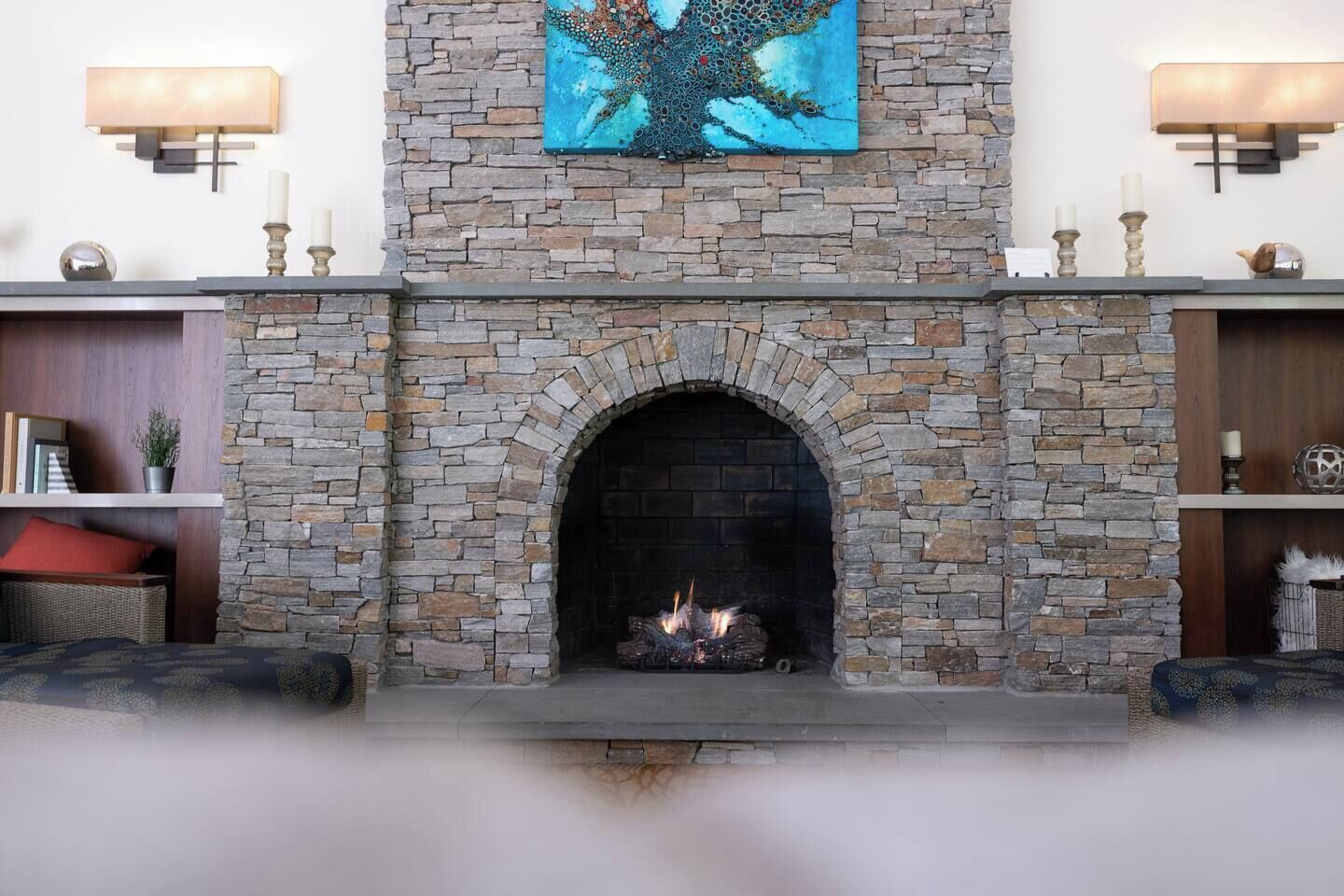 Une cheminée en pierre avec un feu, flanquée d’étagères ornées d’objets décoratifs et surmontée d’un tableau.