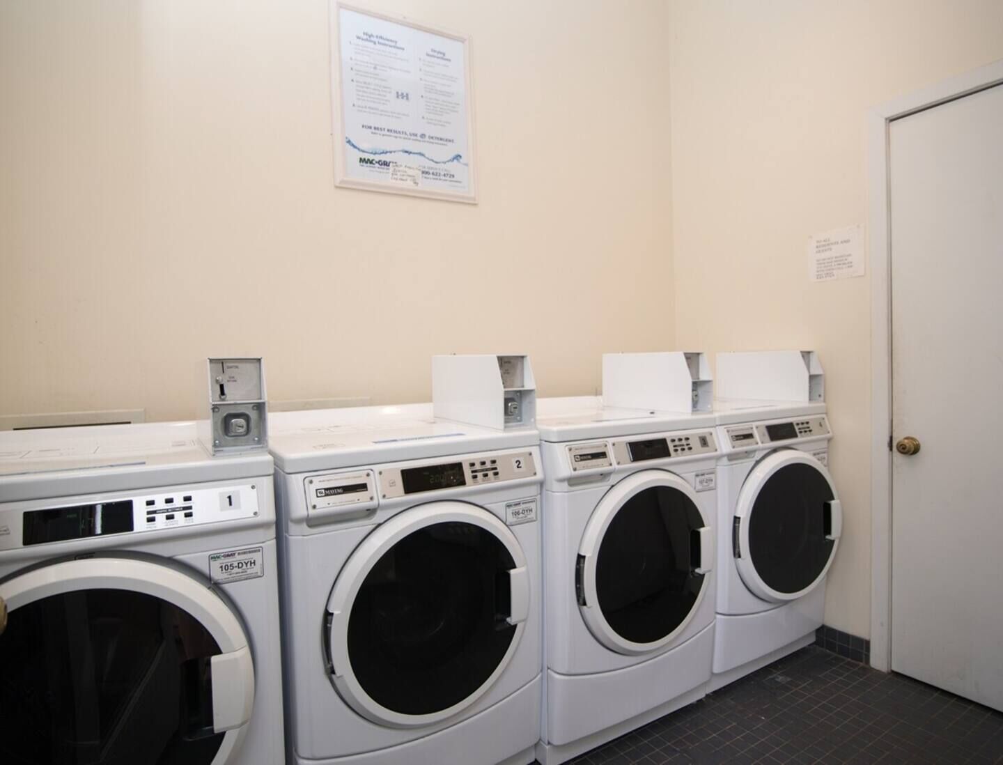 Een wasserette met drie wasmachines en drogers, een bedieningspaneel en een bericht op de muur.
