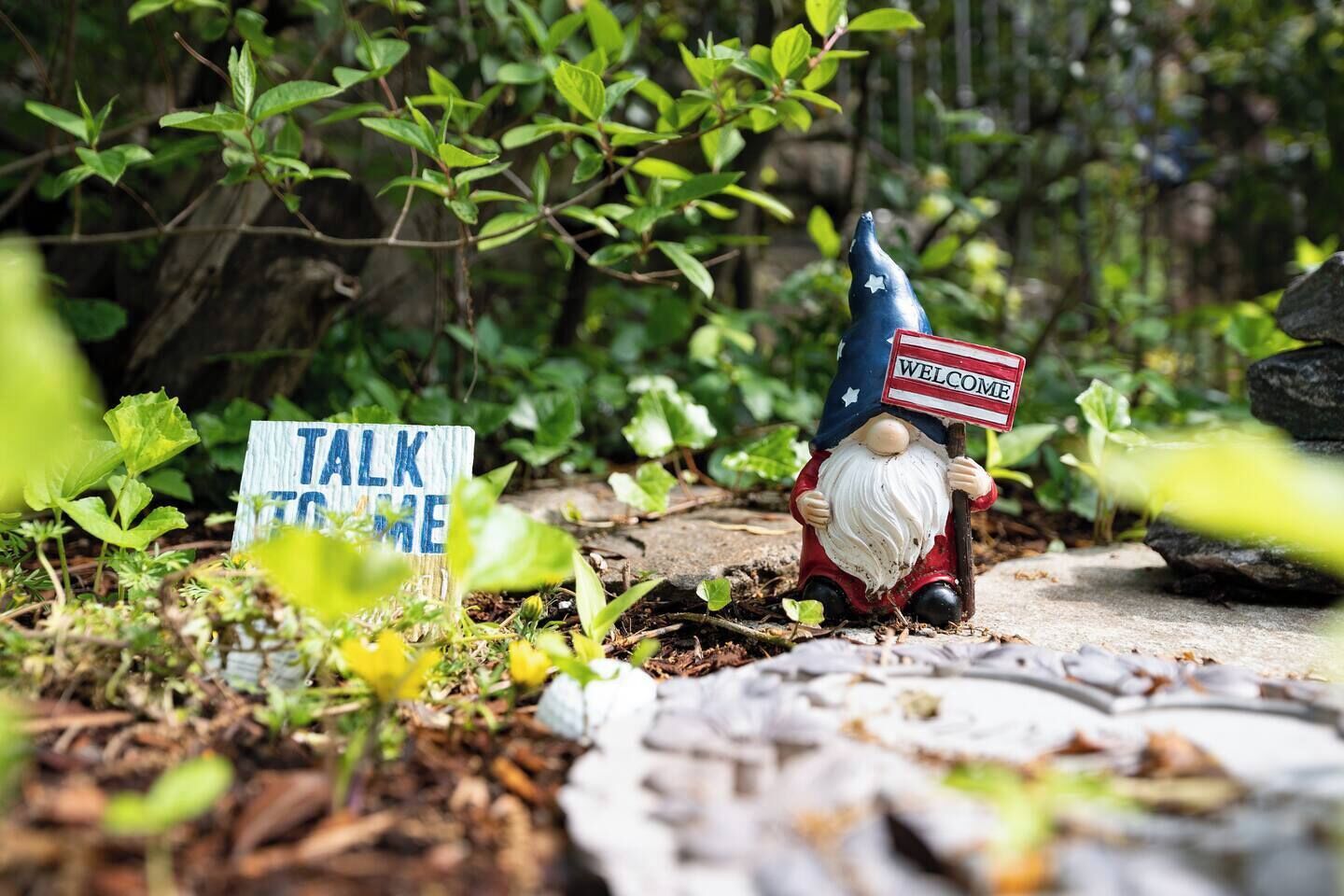 Una statua da giardino raffigura uno gnomo che tiene un cartello con scritto 'WELCOME', con un altro cartello nelle vicinanze che invita a 'TALK TO ME'.