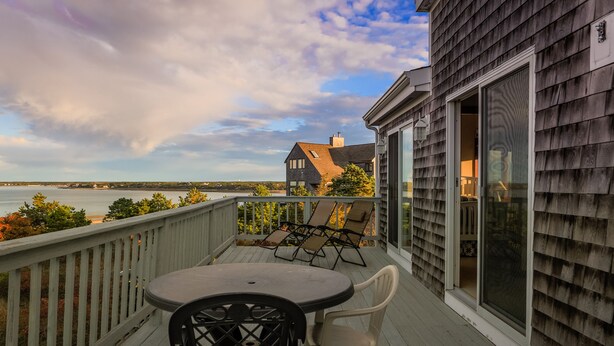 A wooden deck with a table and chairs, overlooking a body of water and a house with a large window.