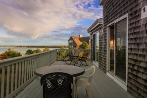 A wooden deck with a table and chairs, overlooking a body of water and a house with a large window. - New for 2026- the perfect family getaway (Wellfleet)