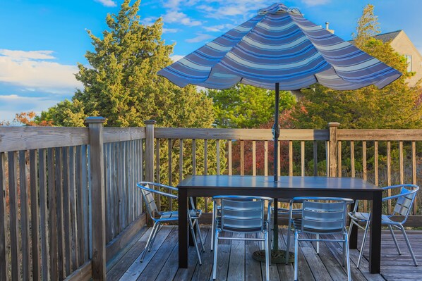 A wooden deck with a table and chairs, an umbrella, and a view of trees and a house in the background. - New for 2026- the perfect family getaway (Wellfleet)