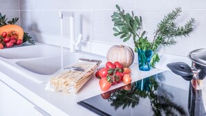 Eine Küchenarbeitsplatte mit Spüle, Schneidebrett, einem Glas Wasser, einer Schüssel mit Tomaten, einem Kürbis, einer Pflanze und einer Packung Nudeln.