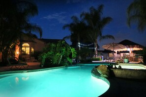A swimming pool with a curved edge, surrounded by palm trees and a building with a thatched roof. - Temecula Wine Country Guest House w/Kitchen - Prime Location (Temecula)