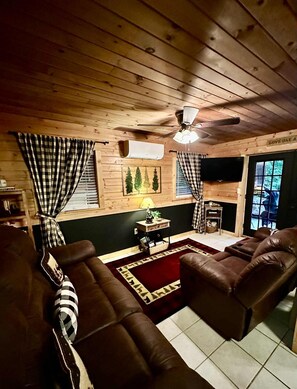 A cozy cabin interior with a fireplace, leather sofas, a television, and a ceiling fan. - Relax at Clear Creek Family Retreat – Cozy Ellijay Cabin by the Water (Ellijay)