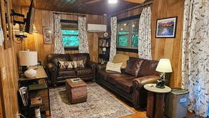 A cozy living room with a brown leather sofa, a small coffee table, a fireplace, and a window with curtains. - Relax at Clear Creek Family Retreat – Cozy Ellijay Cabin by the Water (Ellijay)