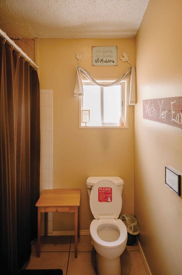 Un baño pequeño con inodoro, un banco de madera, una ventana con cortinas y un cartel que dice 'No Shoes'.