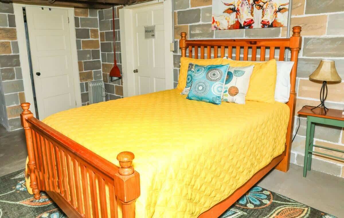 Una cama con estructura de madera y ropa de cama amarilla, una mesita de noche pequeña con una lámpara, una alfombra estampada y una pared con diseño de piedra y un cuadro.