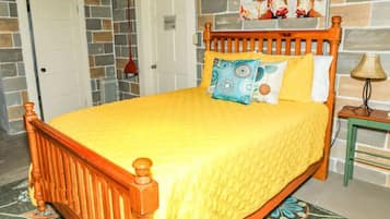 Una cama con estructura de madera y ropa de cama amarilla, una mesita de noche pequeña con una lámpara, una alfombra estampada y una pared con diseño de piedra y un cuadro.